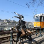 The little Princess statue wearing a mask on the Danube promenade with a tram and the Royal palace in the background