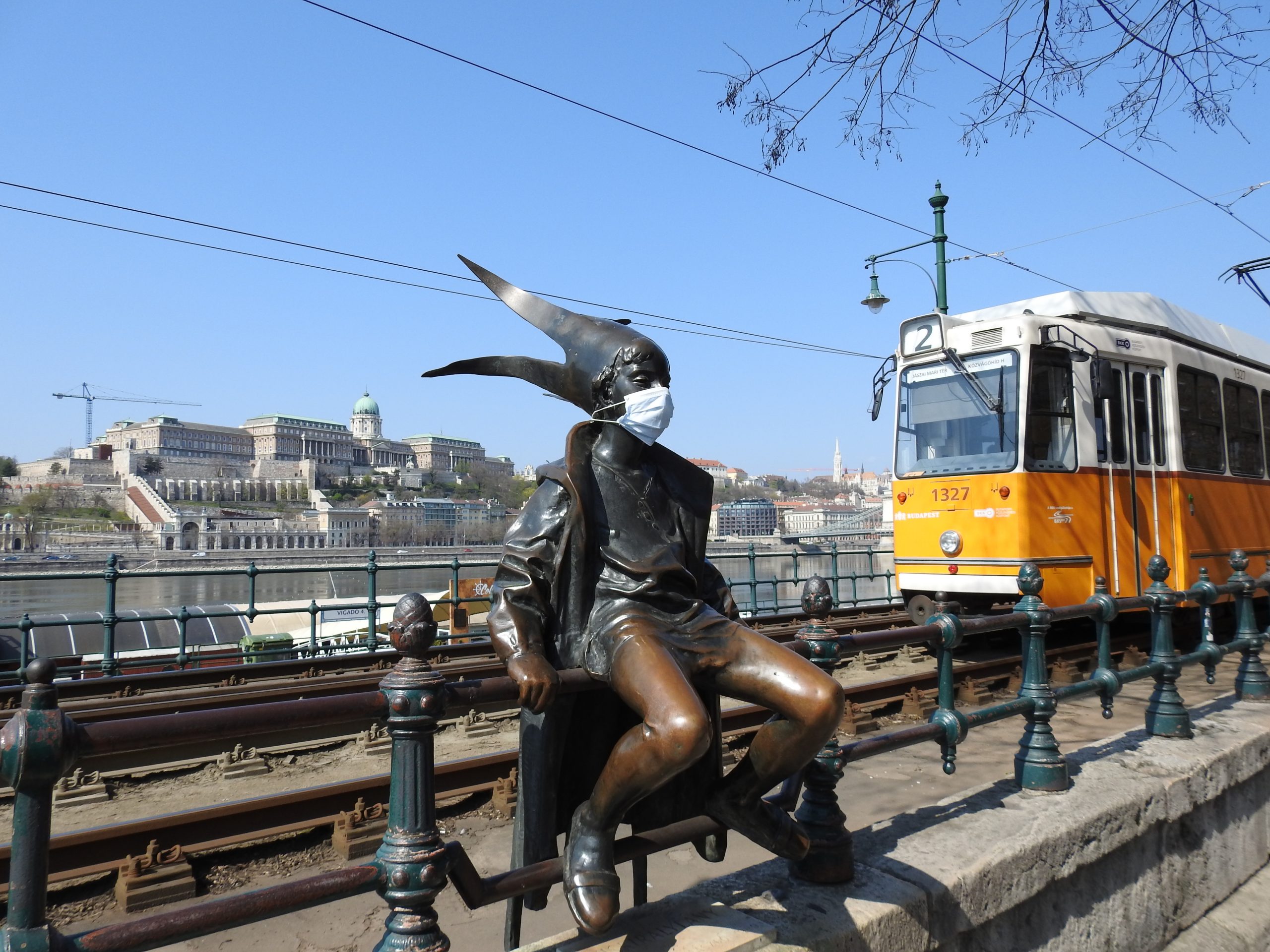 The little Princess statue wearing a mask on the Danube promenade with a tram and the Royal palace in the background